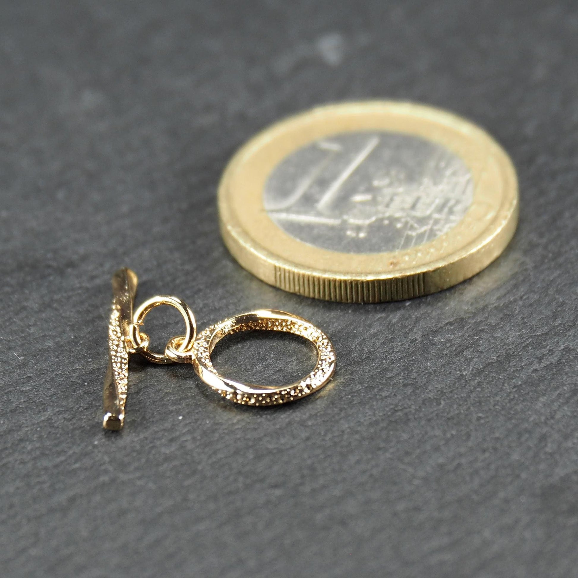 Knebelverschluss, goldfarben, mit runder durchbrochener Öse mit punktierter Struktur und horizontalem Stab, verbunden durch kleine Ringe.