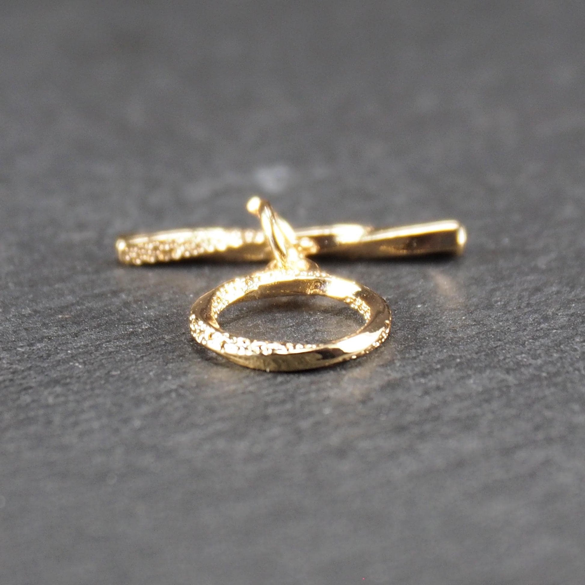 Knebelverschluss, goldfarben, mit runder durchbrochener Öse mit punktierter Struktur und horizontalem Stab, verbunden durch kleine Ringe.