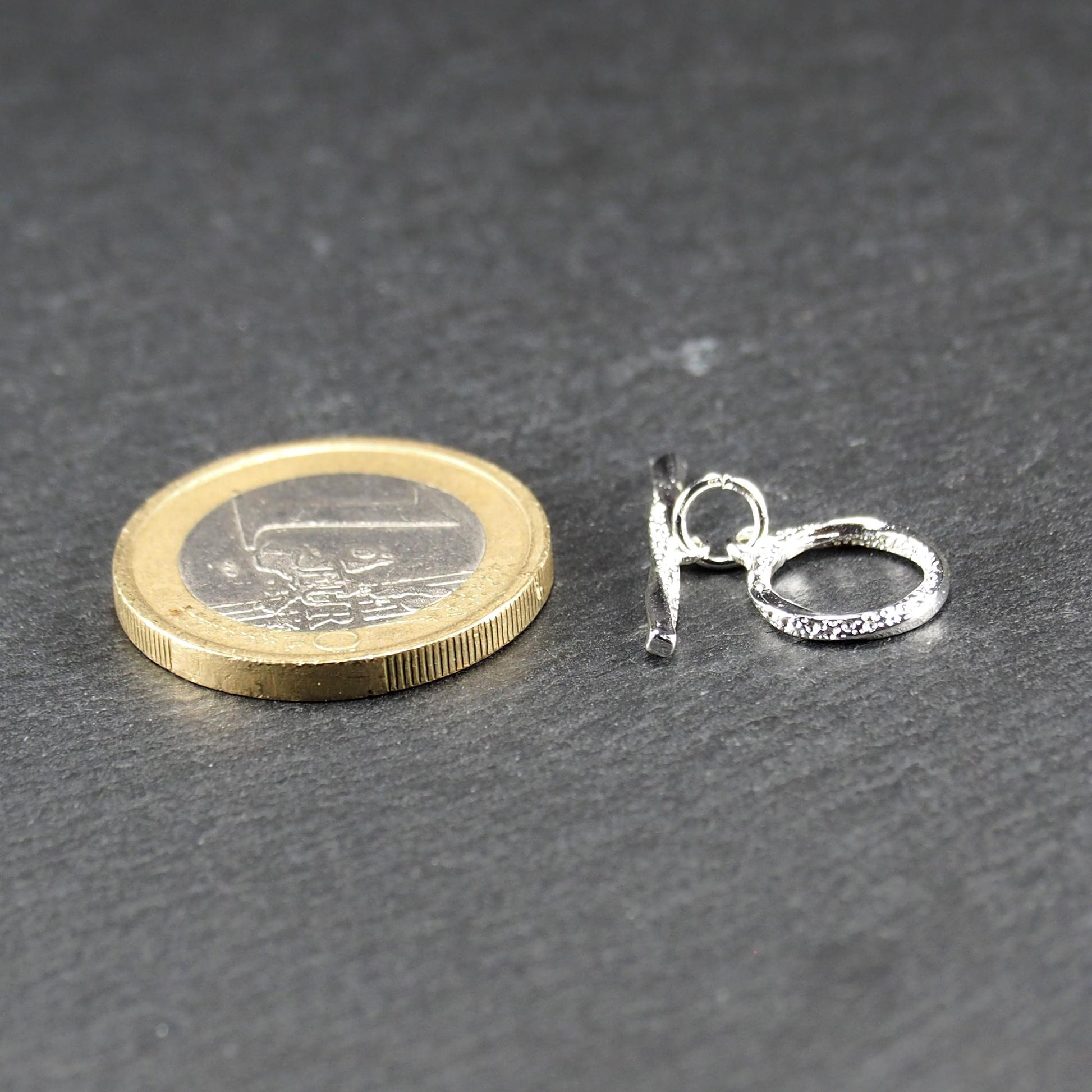 Knebelverschluss (T-Bar) mit silberner, strukturierter Oberfläche, bestehend aus einem horizontalen Stab und einer hängenden, ringförmigen offenen Öse.