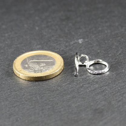 Knebelverschluss (T-Bar) mit silberner, strukturierter Oberfläche, bestehend aus einem horizontalen Stab und einer hängenden, ringförmigen offenen Öse.