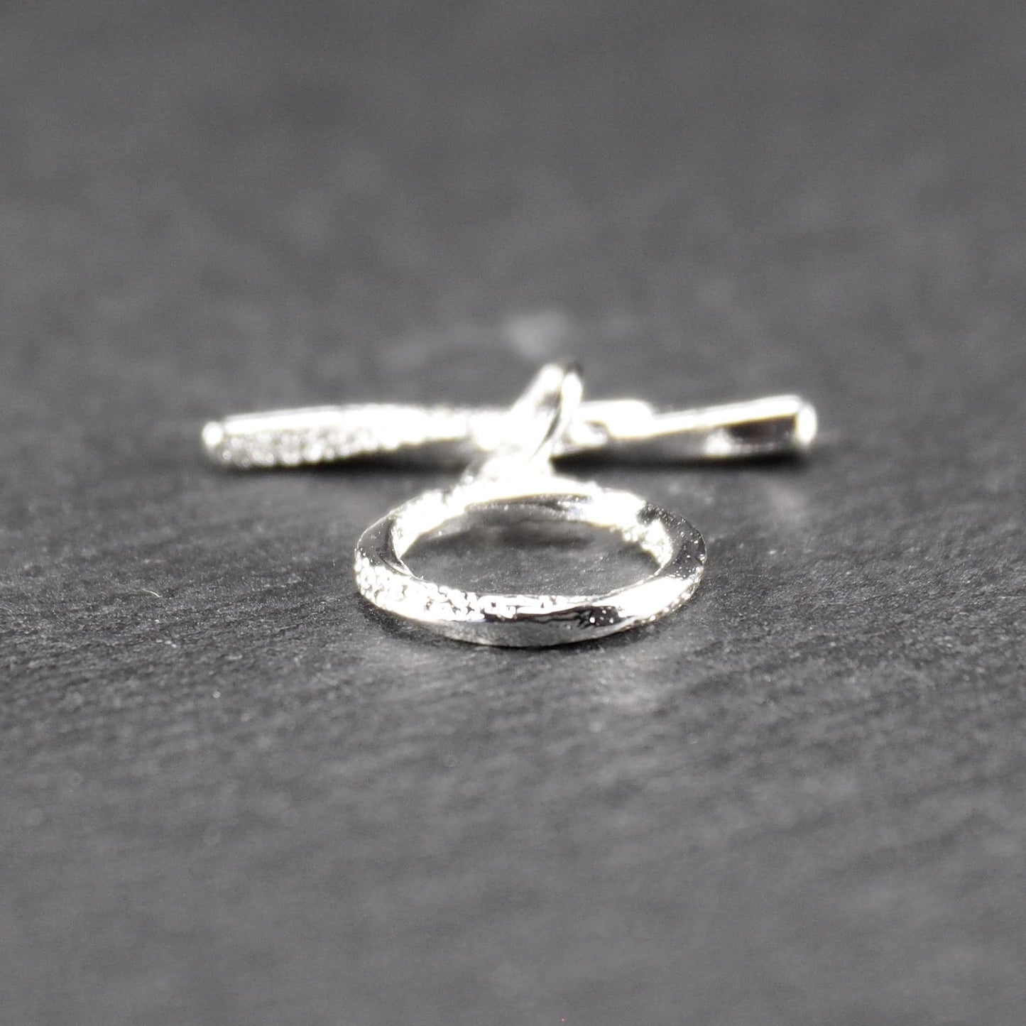 Knebelverschluss (T-Bar) mit silberner, strukturierter Oberfläche, bestehend aus einem horizontalen Stab und einer hängenden, ringförmigen offenen Öse.