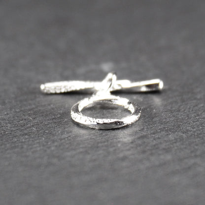 Knebelverschluss (T-Bar) mit silberner, strukturierter Oberfläche, bestehend aus einem horizontalen Stab und einer hängenden, ringförmigen offenen Öse.