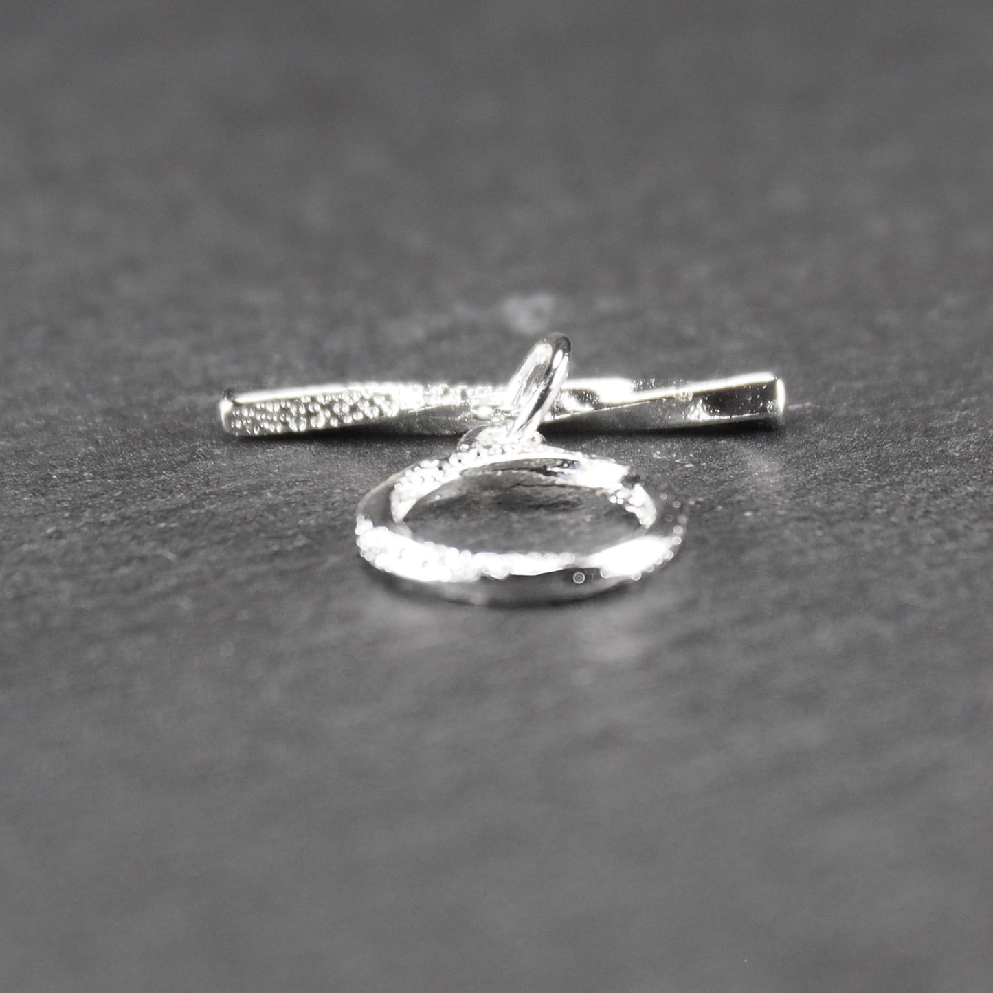 Knebelverschluss (T-Bar) mit silberner, strukturierter Oberfläche, bestehend aus einem horizontalen Stab und einer hängenden, ringförmigen offenen Öse.