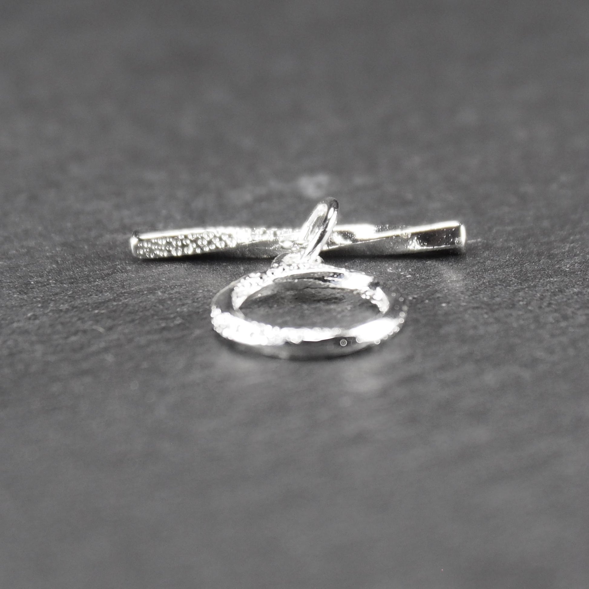 Knebelverschluss (T-Bar) mit silberner, strukturierter Oberfläche, bestehend aus einem horizontalen Stab und einer hängenden, ringförmigen offenen Öse.