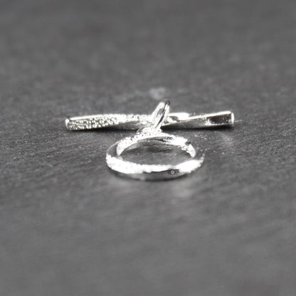 Knebelverschluss (T-Bar) mit silberner, strukturierter Oberfläche, bestehend aus einem horizontalen Stab und einer hängenden, ringförmigen offenen Öse.