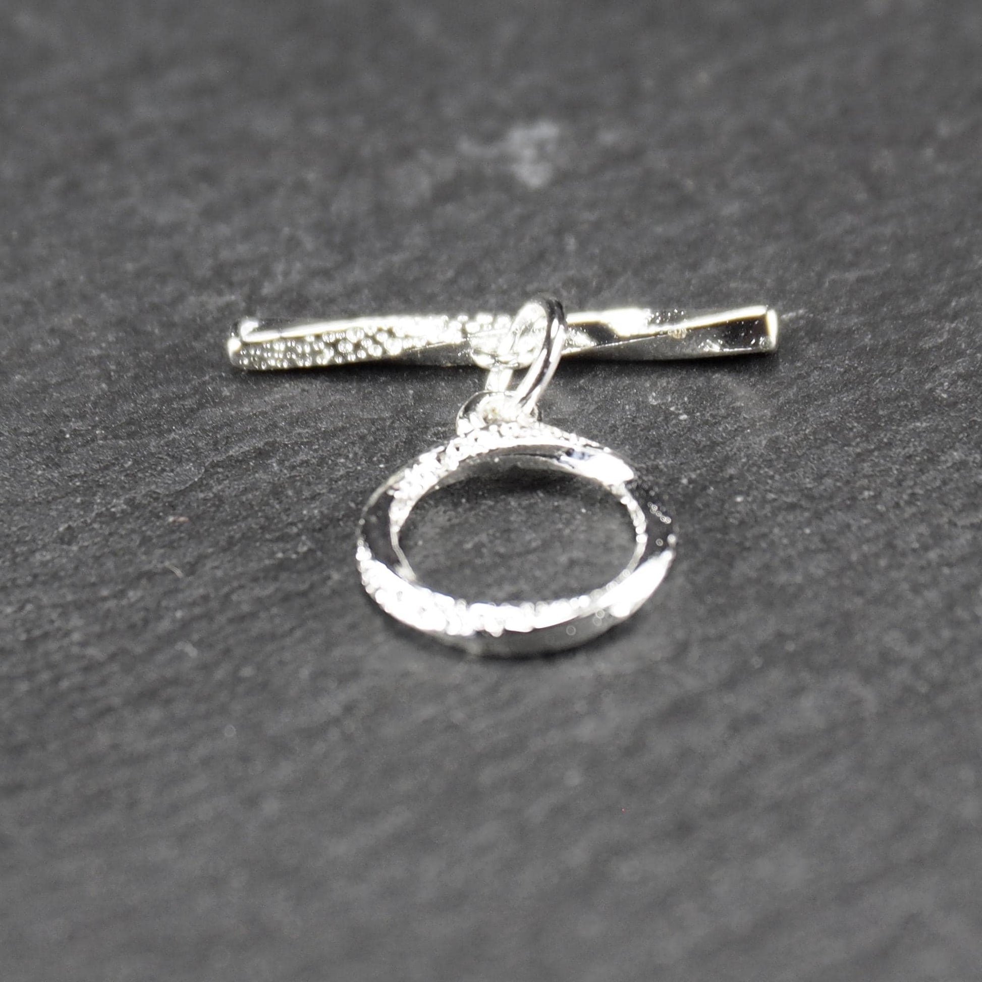 Knebelverschluss (T-Bar) mit silberner, strukturierter Oberfläche, bestehend aus einem horizontalen Stab und einer hängenden, ringförmigen offenen Öse.