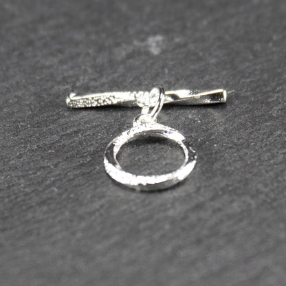 Knebelverschluss (T-Bar) mit silberner, strukturierter Oberfläche, bestehend aus einem horizontalen Stab und einer hängenden, ringförmigen offenen Öse.