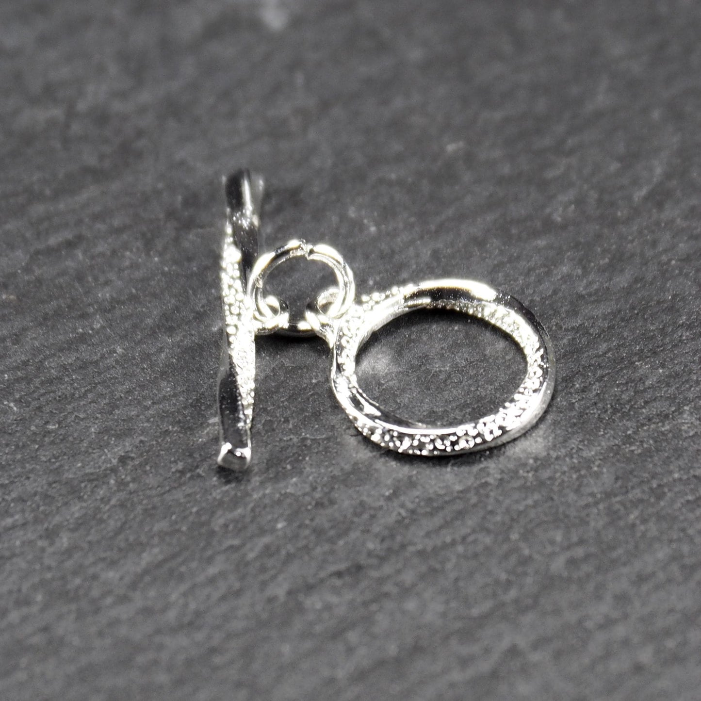 Knebelverschluss (T-Bar) mit silberner, strukturierter Oberfläche, bestehend aus einem horizontalen Stab und einer hängenden, ringförmigen offenen Öse.