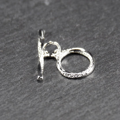 Knebelverschluss (T-Bar) mit silberner, strukturierter Oberfläche, bestehend aus einem horizontalen Stab und einer hängenden, ringförmigen offenen Öse.