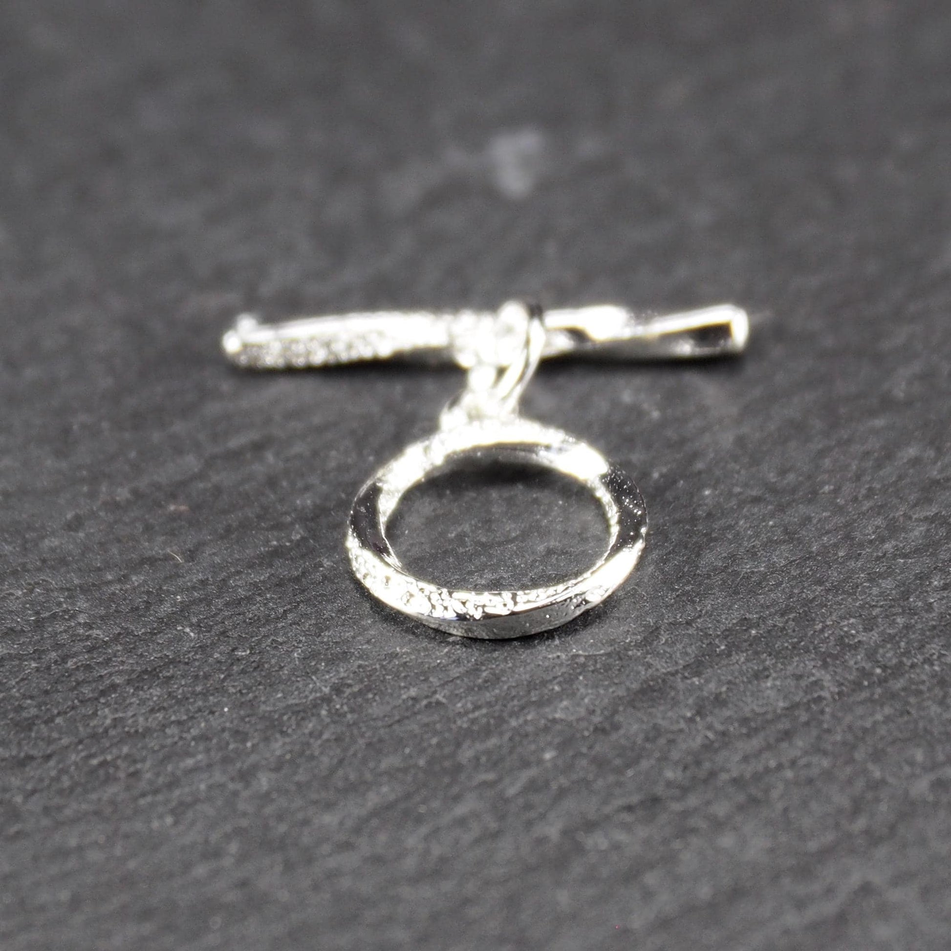 Knebelverschluss (T-Bar) mit silberner, strukturierter Oberfläche, bestehend aus einem horizontalen Stab und einer hängenden, ringförmigen offenen Öse.