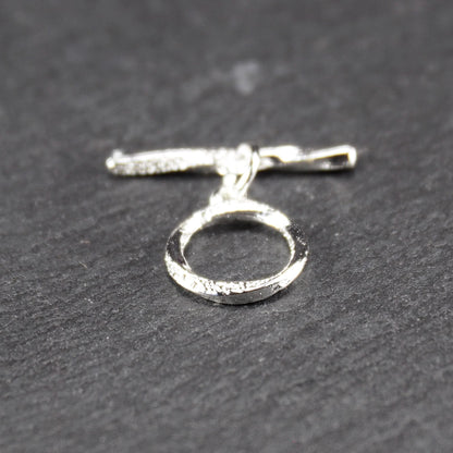 Knebelverschluss (T-Bar) mit silberner, strukturierter Oberfläche, bestehend aus einem horizontalen Stab und einer hängenden, ringförmigen offenen Öse.