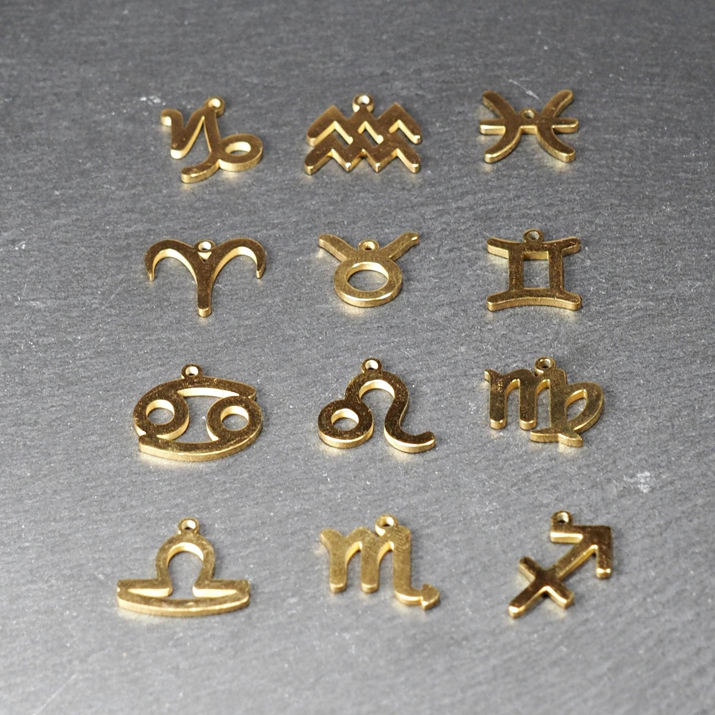 Zwölf flache, vergoldete Sternzeichen-Anhänger aus Metall mit minimalistischer, durchbrochener Kontur und integrierter Öse auf dunklem Hintergrund.