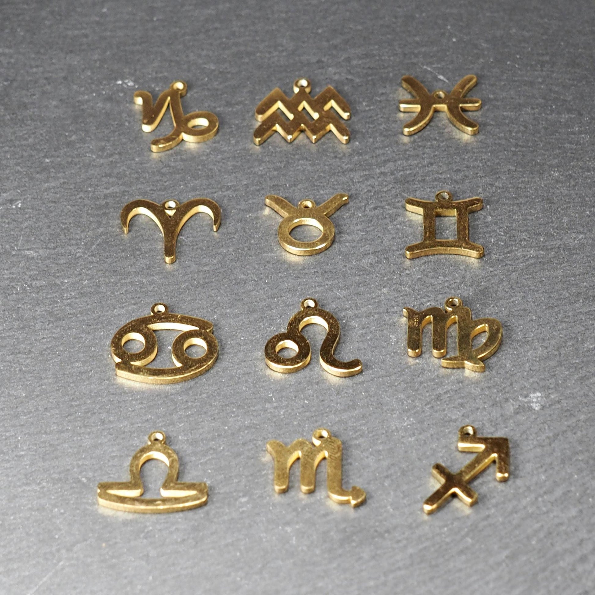 Zwölf flache, vergoldete Sternzeichen-Anhänger aus Metall mit minimalistischer, durchbrochener Kontur und integrierter Öse auf dunklem Hintergrund.