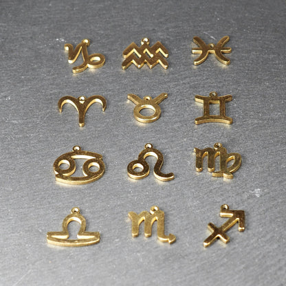 Zwölf flache, vergoldete Sternzeichen-Anhänger aus Metall mit minimalistischer, durchbrochener Kontur und integrierter Öse auf dunklem Hintergrund.