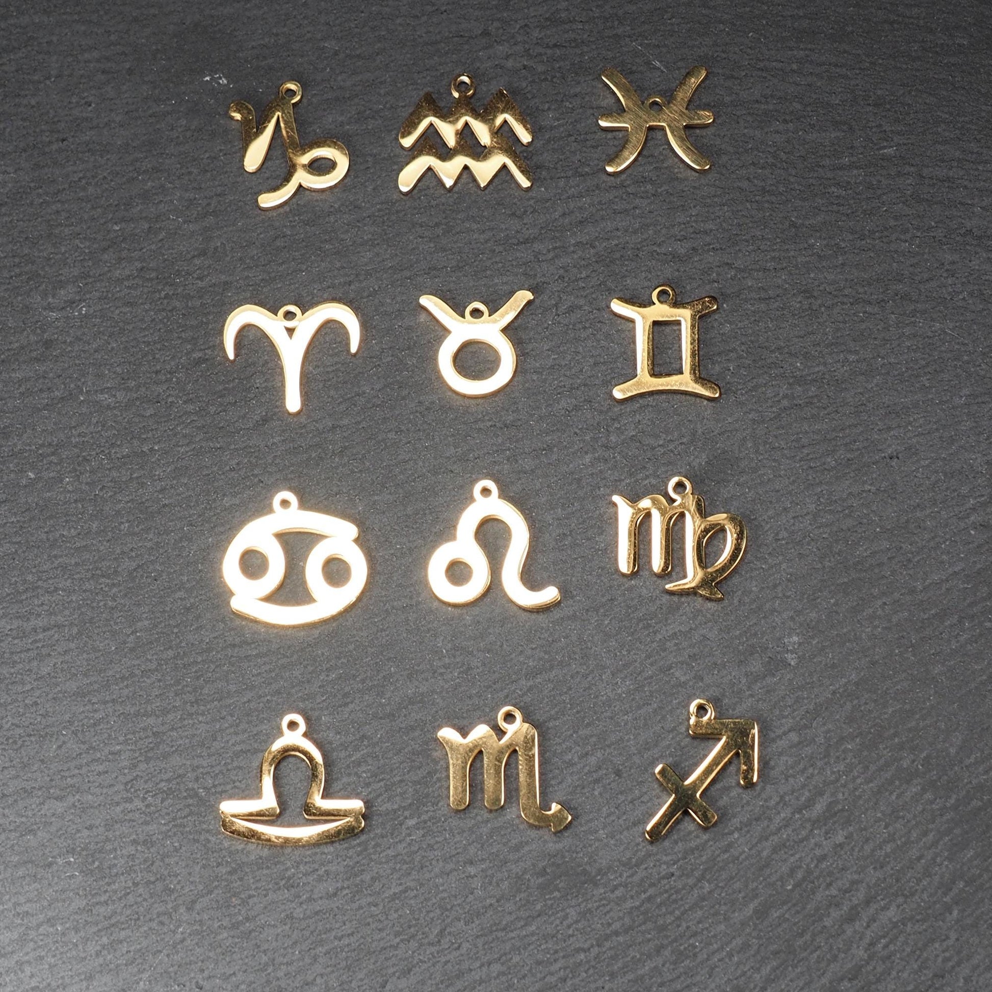 Zwölf flache, vergoldete Sternzeichen-Anhänger aus Metall mit minimalistischer, durchbrochener Kontur und integrierter Öse auf dunklem Hintergrund.