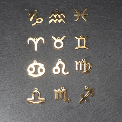 Zwölf flache, vergoldete Sternzeichen-Anhänger aus Metall mit minimalistischer, durchbrochener Kontur und integrierter Öse auf dunklem Hintergrund.