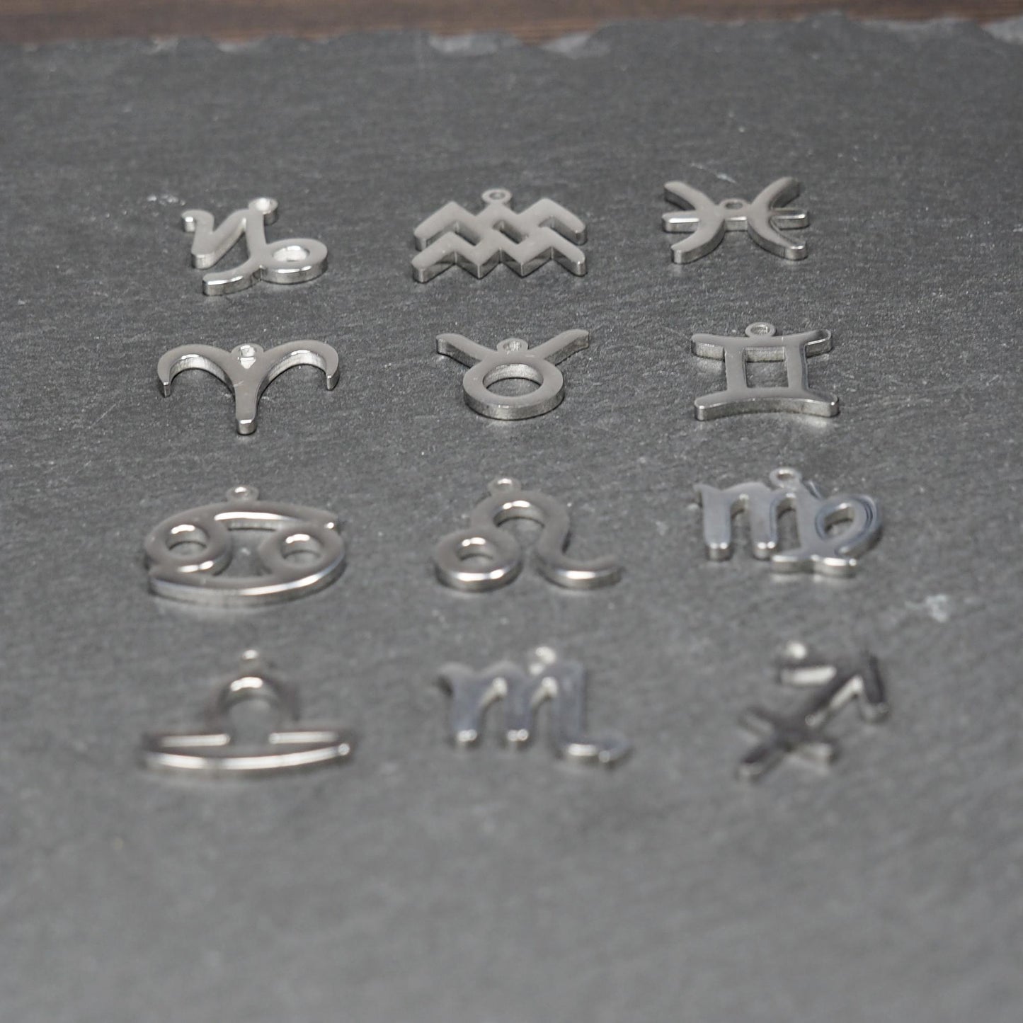 Zwölf flache silberfarbene Edelstahl-Anhänger als durchbrochene Sternzeichensymbole mit glänzender Oberfläche und je einer Öse auf dunklem Hintergrund.
