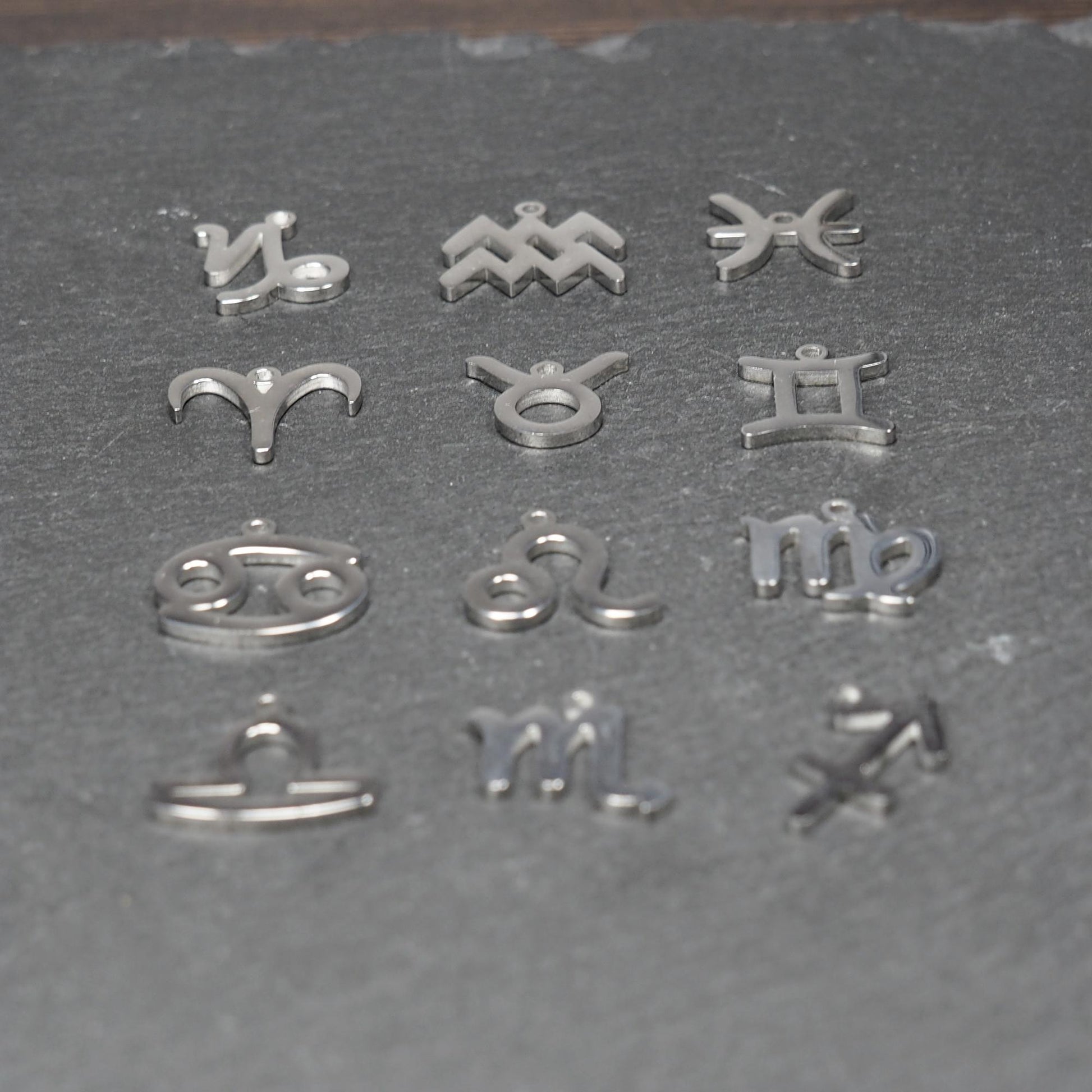 Zwölf flache silberfarbene Edelstahl-Anhänger als durchbrochene Sternzeichensymbole mit glänzender Oberfläche und je einer Öse auf dunklem Hintergrund.