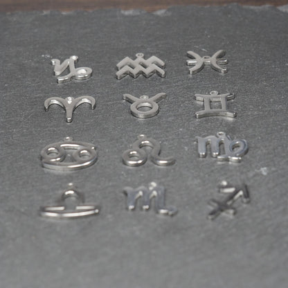 Zwölf flache silberfarbene Edelstahl-Anhänger als durchbrochene Sternzeichensymbole mit glänzender Oberfläche und je einer Öse auf dunklem Hintergrund.
