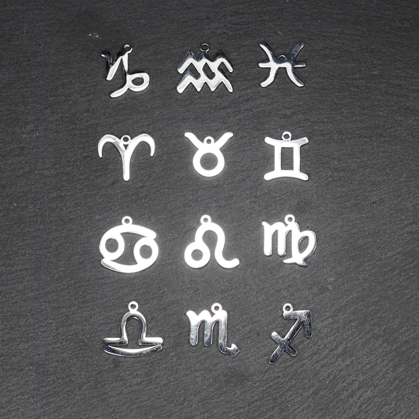 Zwölf flache silberfarbene Edelstahl-Anhänger als durchbrochene Sternzeichensymbole mit glänzender Oberfläche und je einer Öse auf dunklem Hintergrund.
