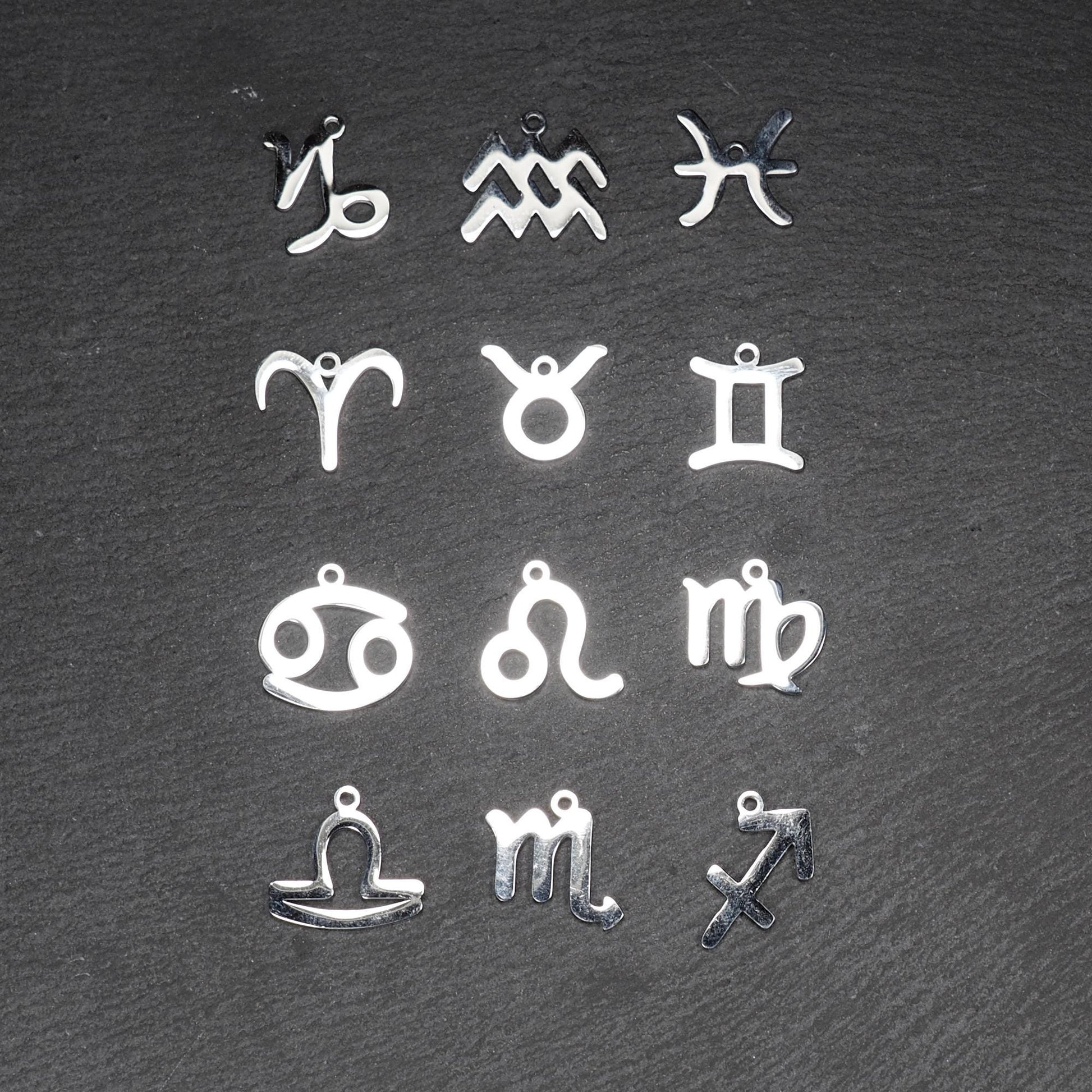 Zwölf flache silberfarbene Edelstahl-Anhänger als durchbrochene Sternzeichensymbole mit glänzender Oberfläche und je einer Öse auf dunklem Hintergrund.