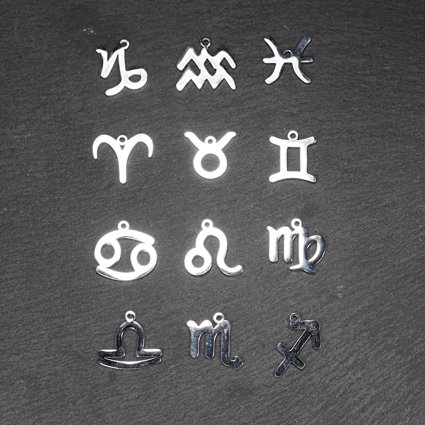 Zwölf flache silberfarbene Edelstahl-Anhänger als durchbrochene Sternzeichensymbole mit glänzender Oberfläche und je einer Öse auf dunklem Hintergrund.