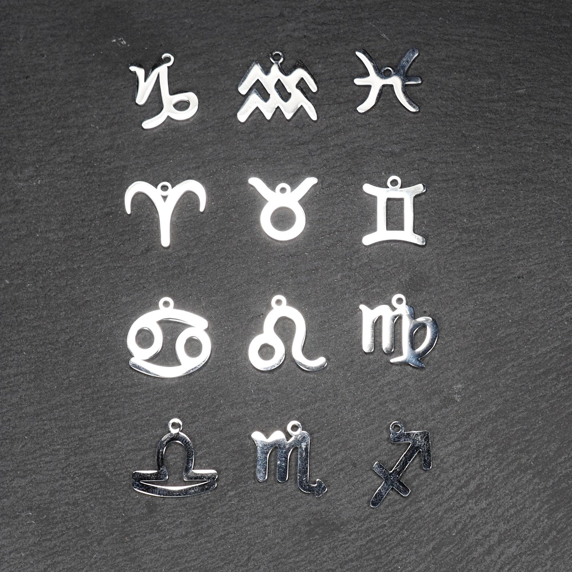 Zwölf flache silberfarbene Edelstahl-Anhänger als durchbrochene Sternzeichensymbole mit glänzender Oberfläche und je einer Öse auf dunklem Hintergrund.