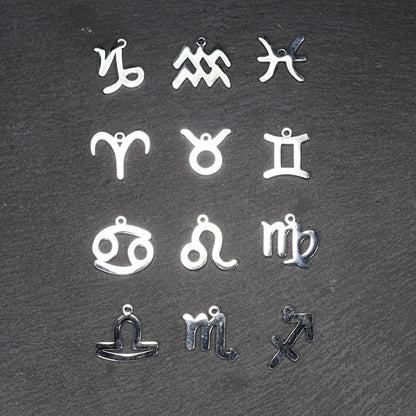 Zwölf flache silberfarbene Edelstahl-Anhänger als durchbrochene Sternzeichensymbole mit glänzender Oberfläche und je einer Öse auf dunklem Hintergrund.