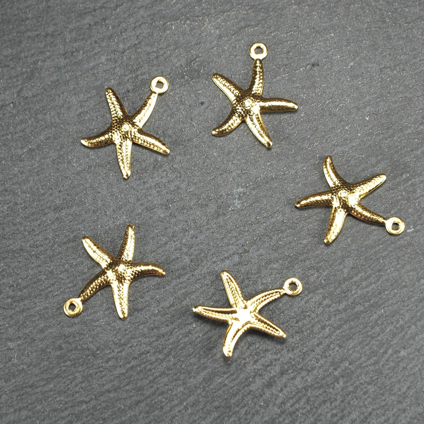 Fünf goldfarbene, flache Seestern-Anhänger mit reliefartiger, strukturierter und glänzender Oberfläche, jeweils mit kleiner runder Öse zum Aufhängen.