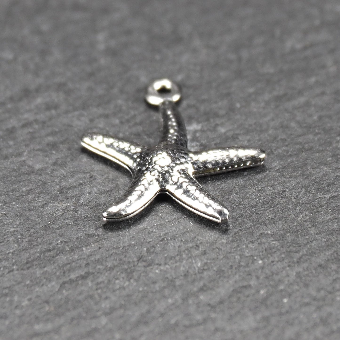 Seestern-Anhänger aus silberfarbenem Metall mit reliefartiger, strukturierter glänzender Oberfläche, fünf Armen und kleiner runder Öse oben.