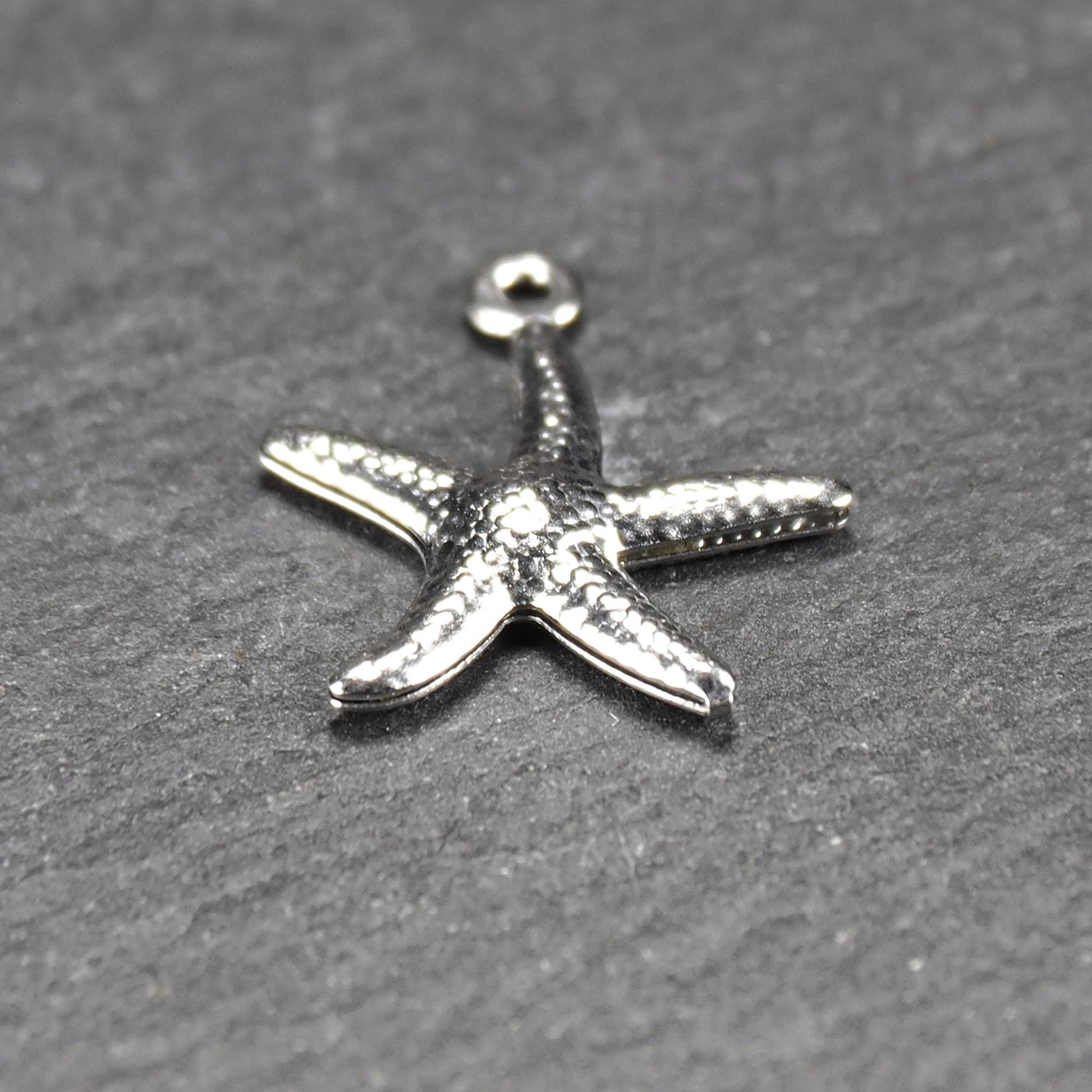 Seestern-Anhänger aus silberfarbenem Metall mit reliefartiger, strukturierter glänzender Oberfläche, fünf Armen und kleiner runder Öse oben.
