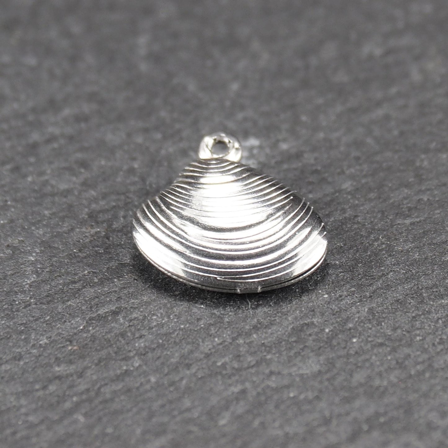 Muschelförmiger silberfarbener Anhänger mit polierter, glänzender Oberfläche, konzentrisch geriffelten Rillen und integrierter runder Öse oben.