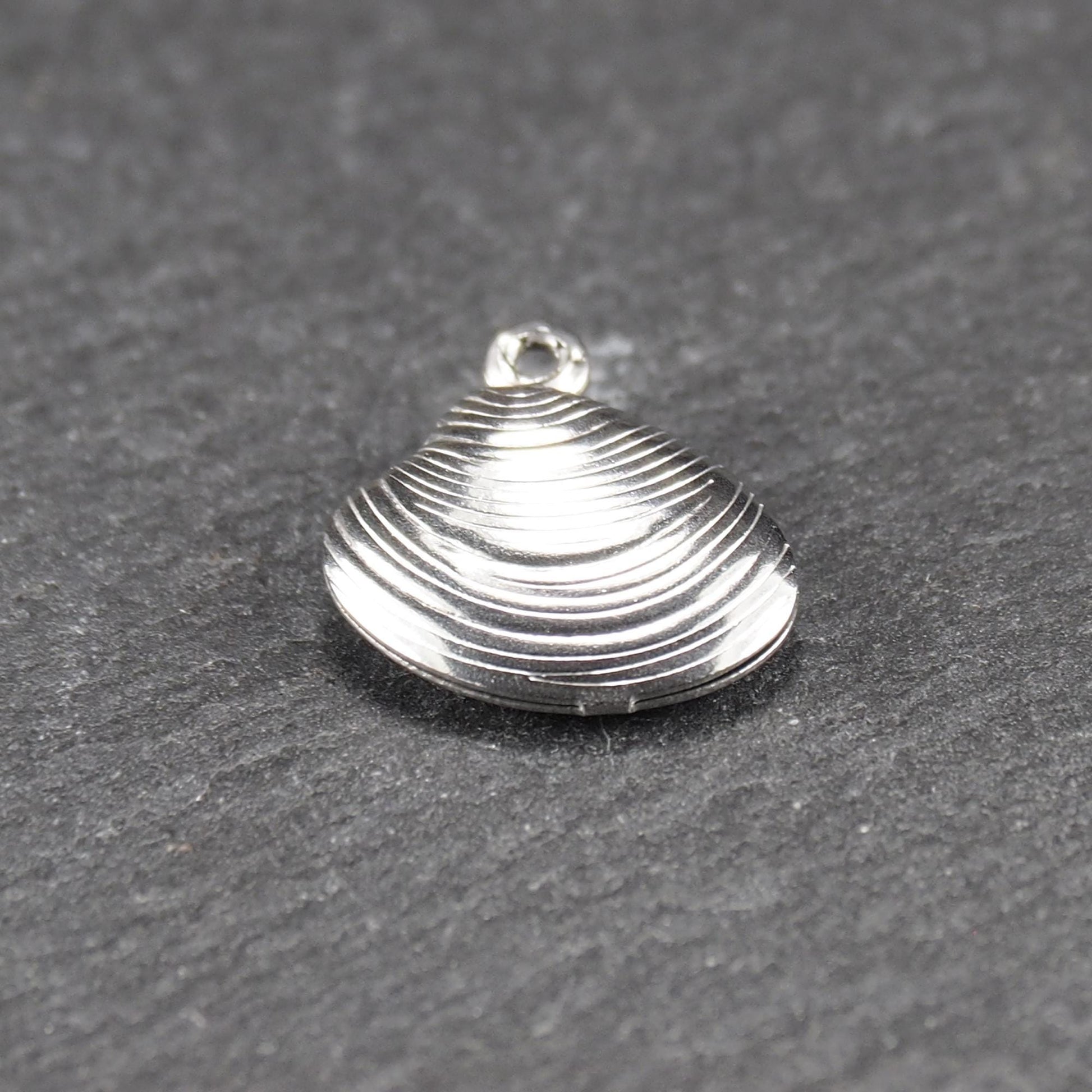 Muschelförmiger silberfarbener Anhänger mit polierter, glänzender Oberfläche, konzentrisch geriffelten Rillen und integrierter runder Öse oben.