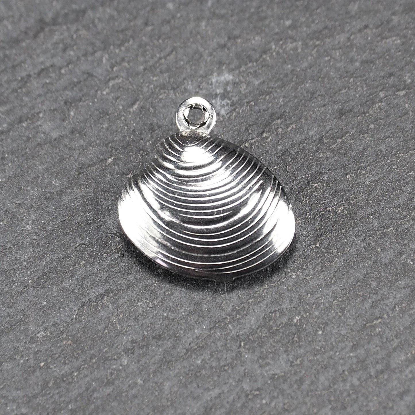 Muschelförmiger silberfarbener Anhänger mit polierter, glänzender Oberfläche, konzentrisch geriffelten Rillen und integrierter runder Öse oben.
