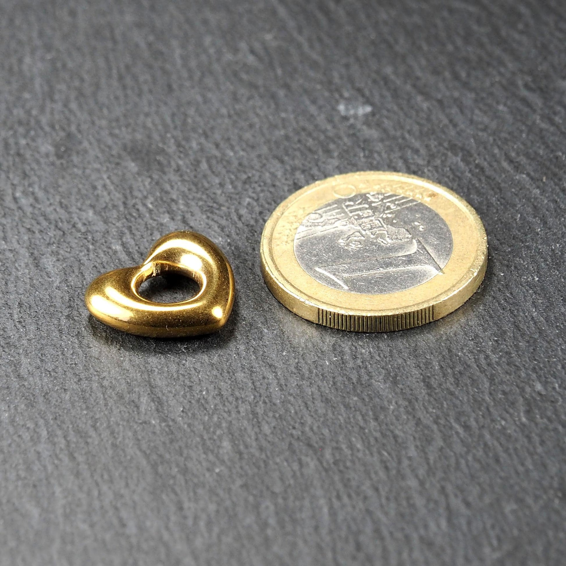 Herzförmiger, goldglänzender Anhänger mit offenem, durchbrochenem Zentrum und rundem Fädelloch, daneben 1-Euro-Münze als Größenvergleich.