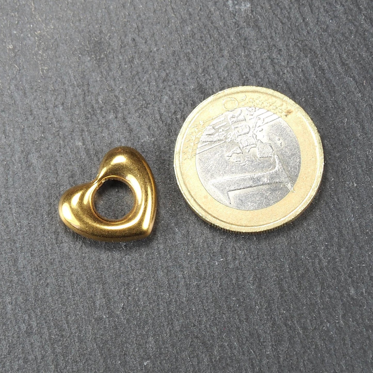 Herzförmiger, goldglänzender Anhänger mit offenem, durchbrochenem Zentrum und rundem Fädelloch, daneben 1-Euro-Münze als Größenvergleich.