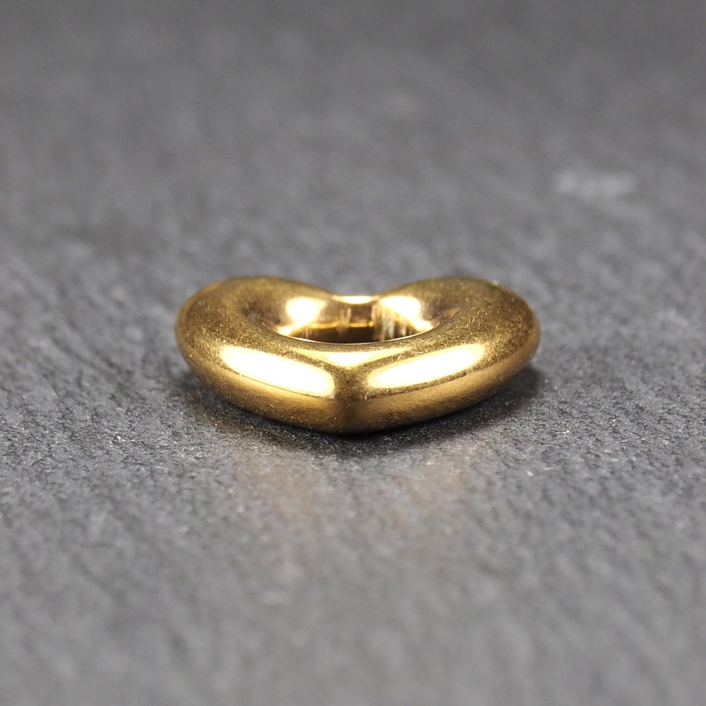 Herzförmiger, goldfarbener Anhänger mit hochglänzender, glatter Oberfläche, rundem durchbrochenem Zentrum und sichtbarer Bohrung.