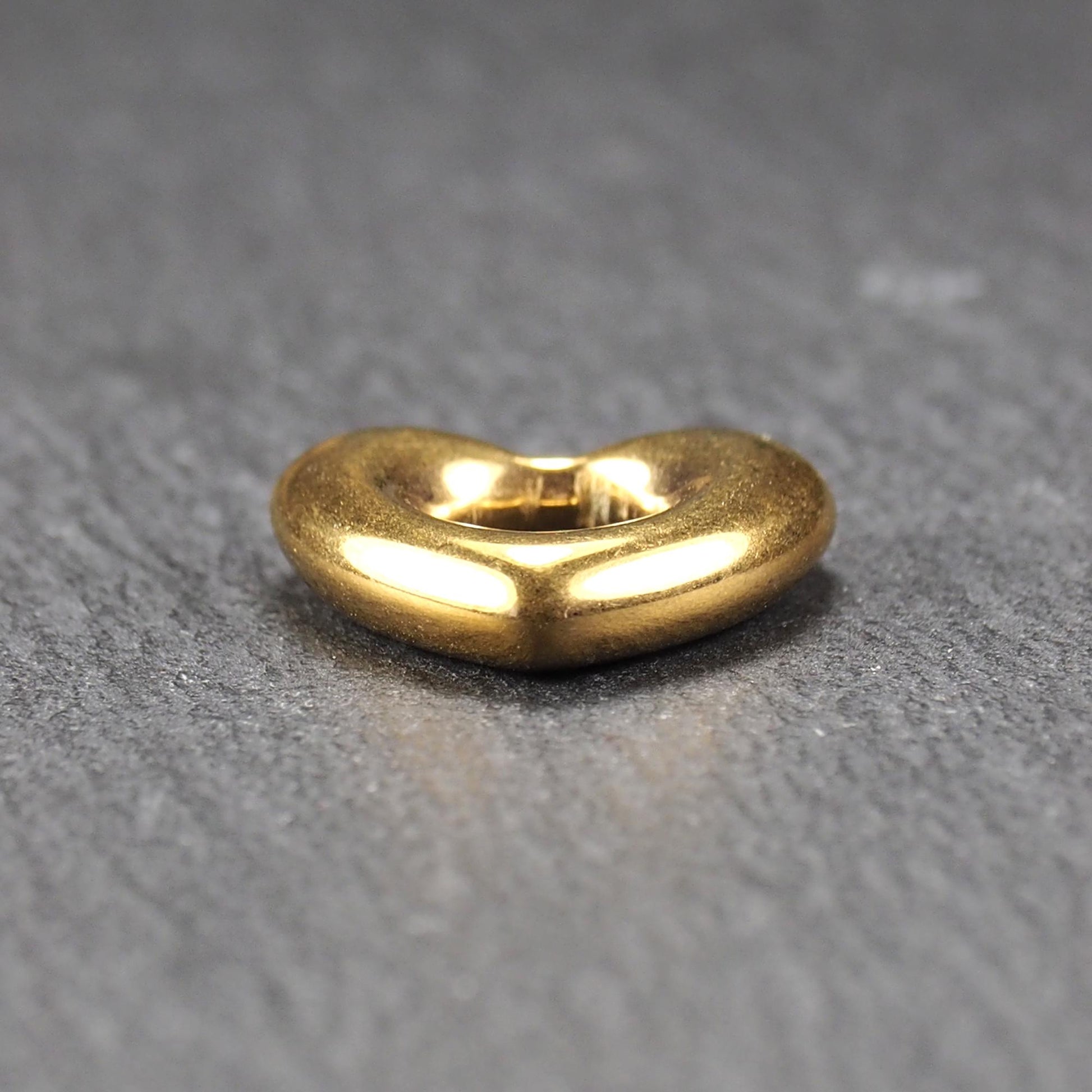 Herzförmiger, goldfarbener Anhänger mit hochglänzender, glatter Oberfläche, rundem durchbrochenem Zentrum und sichtbarer Bohrung.