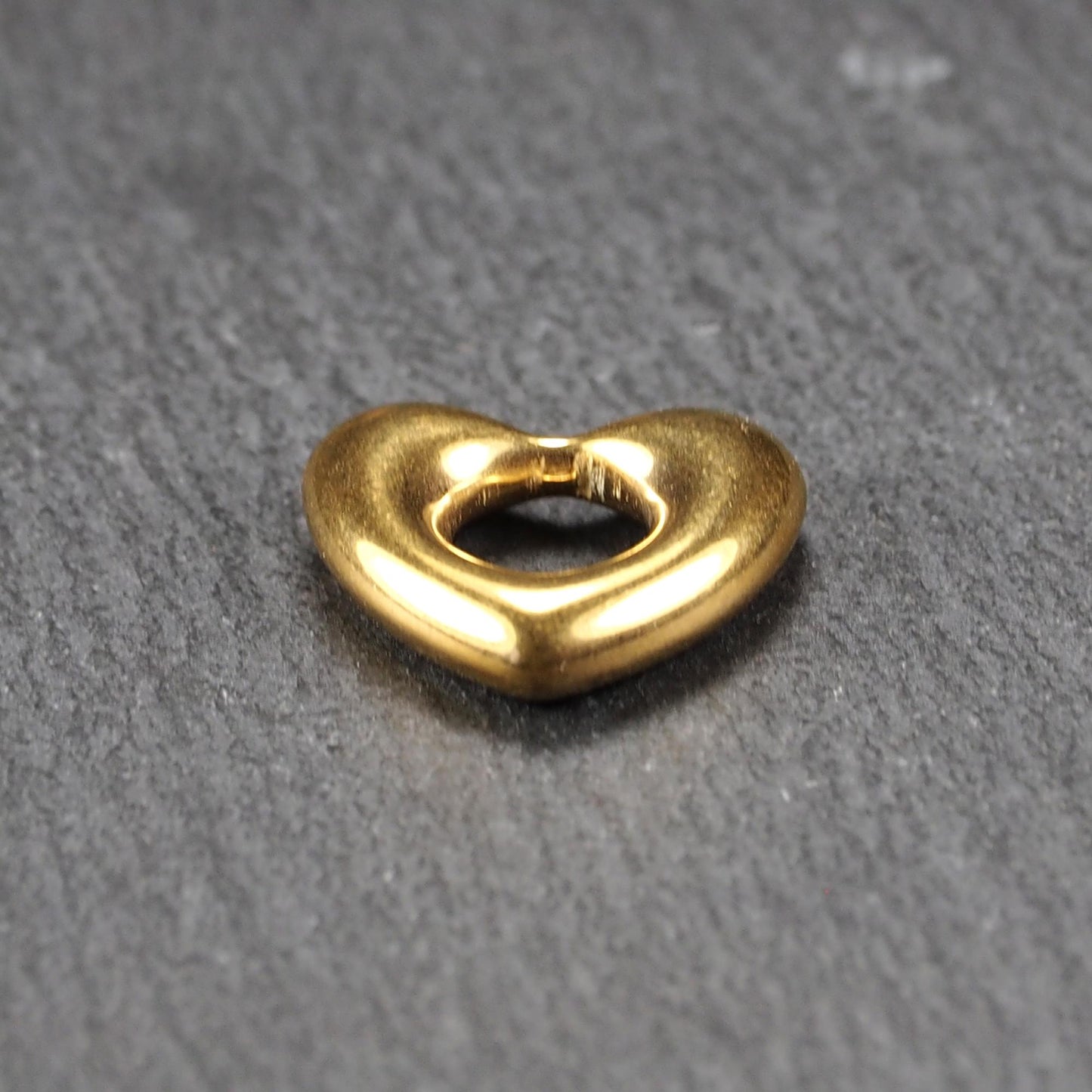 Herzförmiger, goldfarbener Anhänger mit hochglänzender, glatter Oberfläche, rundem durchbrochenem Zentrum und sichtbarer Bohrung.