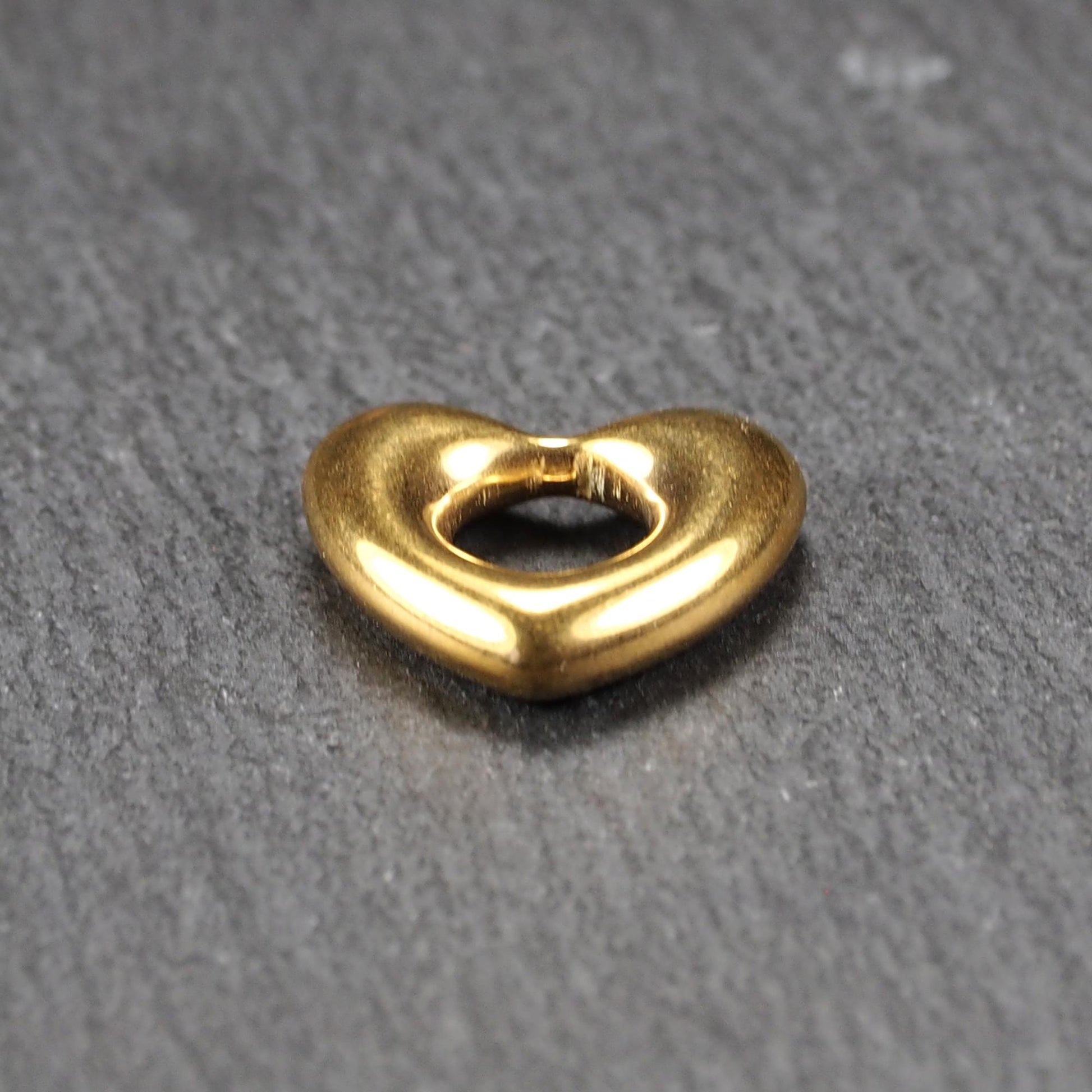 Herzförmiger, goldfarbener Anhänger mit hochglänzender, glatter Oberfläche, rundem durchbrochenem Zentrum und sichtbarer Bohrung.