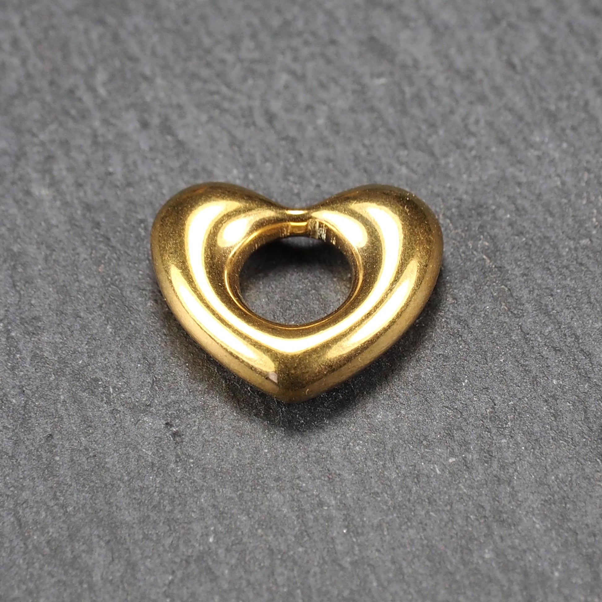 Herzförmiger, goldfarbener Anhänger mit hochglänzender, glatter Oberfläche, rundem durchbrochenem Zentrum und sichtbarer Bohrung.