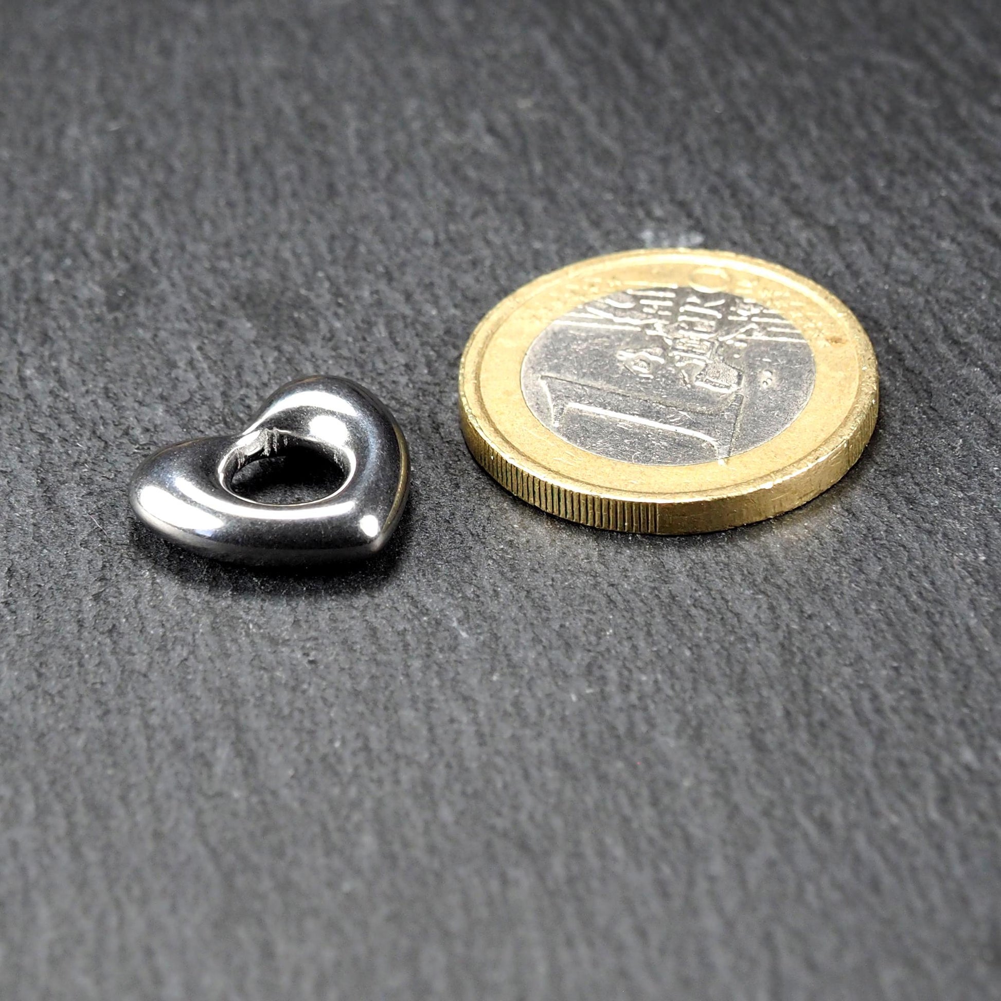 Herzanhänger aus glänzend poliertem Edelstahl mit offenem, durchbrochenem Innenfeld und abgerundeten Kanten, neben einer 1-Euro-Münze.