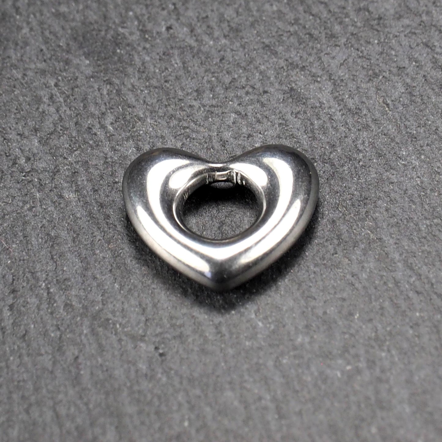 Herzanhänger, glänzend poliertes silbernes Metall mit rundem, durchbrochenem Innenfeld und glatten, abgerundeten Kanten auf dunklem Hintergrund.