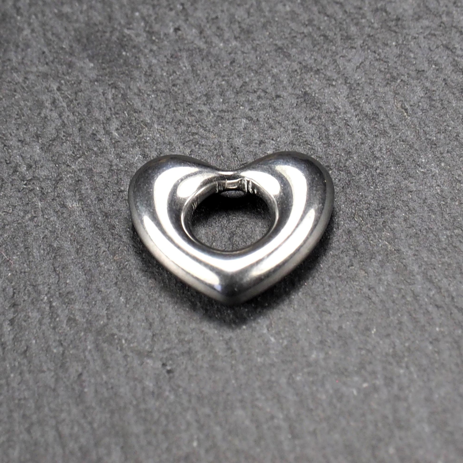 Herzanhänger, glänzend poliertes silbernes Metall mit rundem, durchbrochenem Innenfeld und glatten, abgerundeten Kanten auf dunklem Hintergrund.