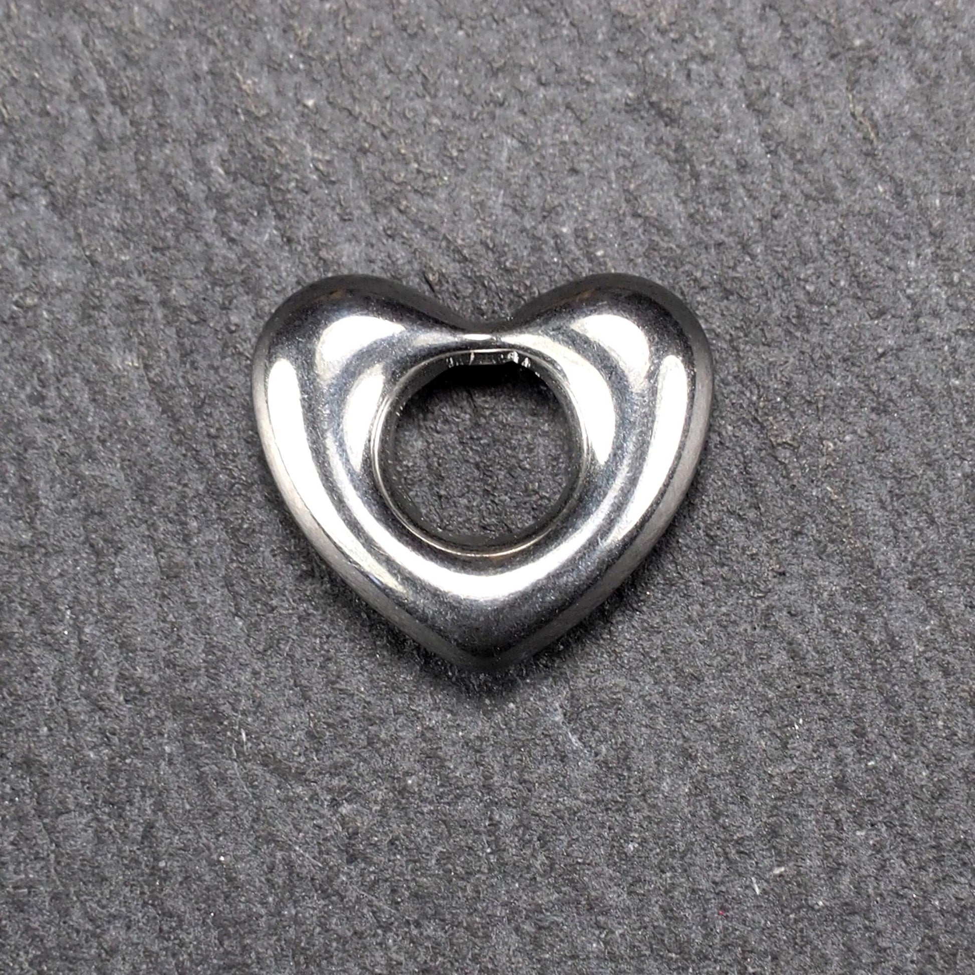 Herzanhänger, glänzend poliertes silbernes Metall mit rundem, durchbrochenem Innenfeld und glatten, abgerundeten Kanten auf dunklem Hintergrund.