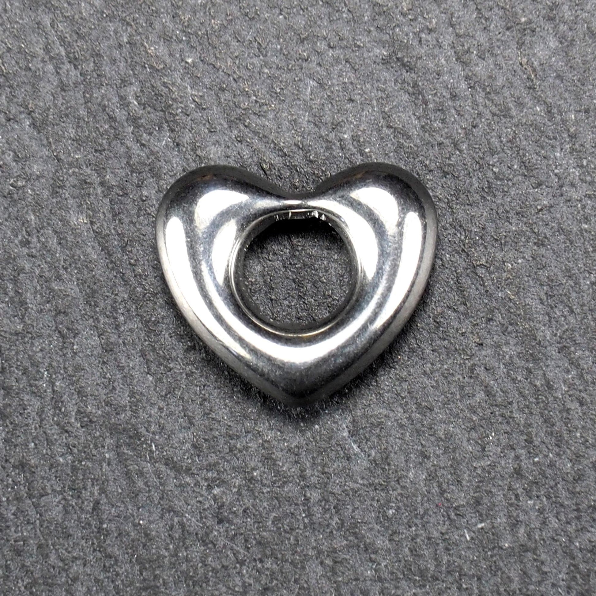 Herzanhänger, glänzend poliertes silbernes Metall mit rundem, durchbrochenem Innenfeld und glatten, abgerundeten Kanten auf dunklem Hintergrund.