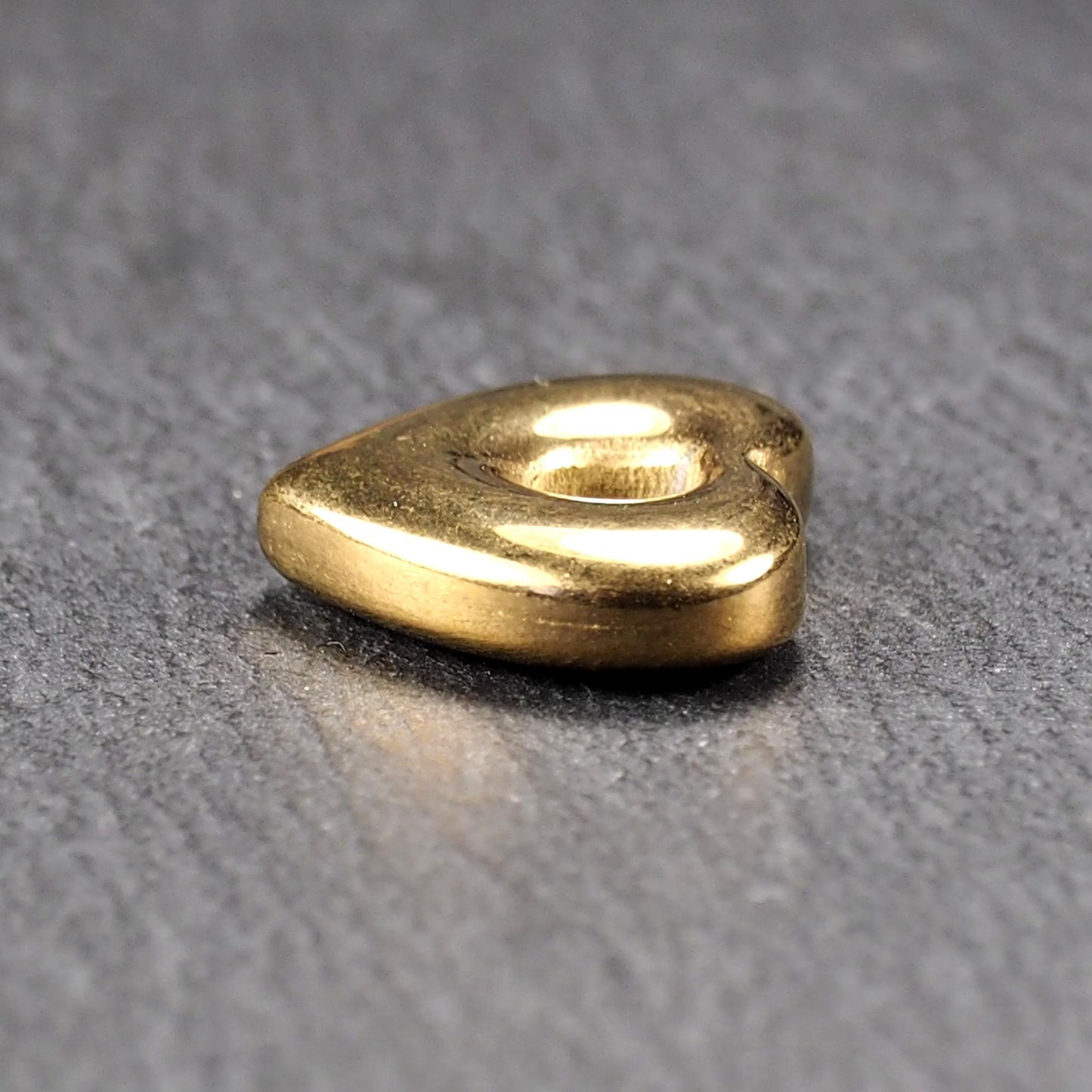 Goldfarbenes, hochglänzendes, abgerundetes Herz mit rundem mittigem Loch, glatte Oberfläche, auf dunkel strukturiertem Untergrund.