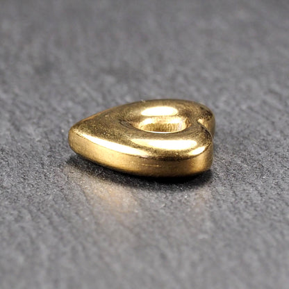 Goldfarbenes, hochglänzendes, abgerundetes Herz mit rundem mittigem Loch, glatte Oberfläche, auf dunkel strukturiertem Untergrund.