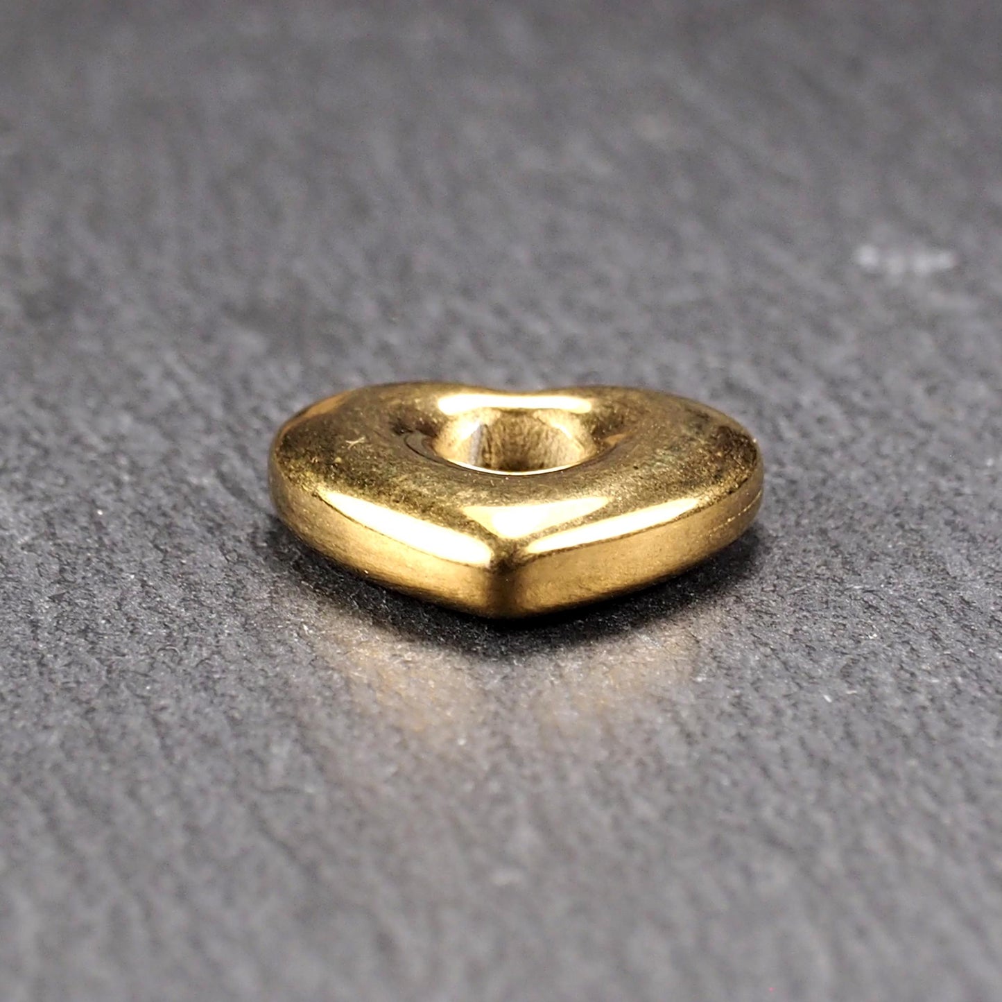 Goldfarbenes, hochglänzendes, abgerundetes Herz mit rundem mittigem Loch, glatte Oberfläche, auf dunkel strukturiertem Untergrund.