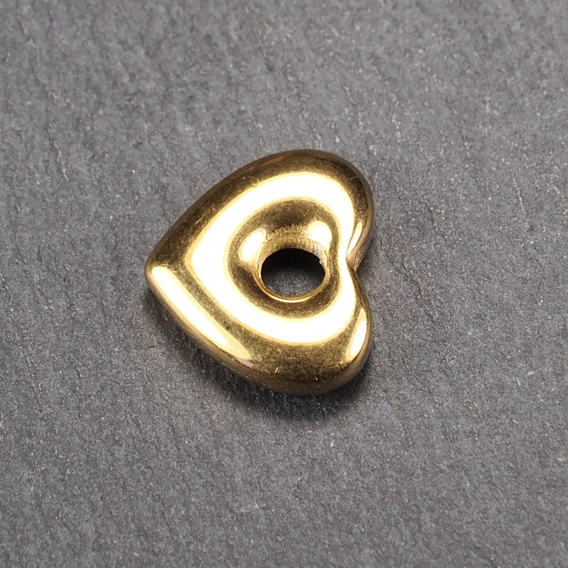 Goldfarbenes, hochglänzendes, abgerundetes Herz mit rundem mittigem Loch, glatte Oberfläche, auf dunkel strukturiertem Untergrund.